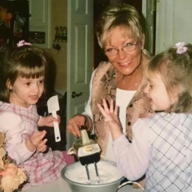 Two young girls sit on a kitchen counter, one holding a spatula, while their grandmother in between them mixes a bowl of white frosting with a hand mixer.