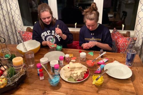 Twin sisters sit at a kitchen table, decorated cookies with various multicolored sprinkles and frosting.