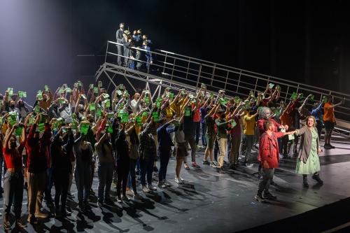 Dozens of performers on stage, all but a couple in front holding up green slips of paper. A ramp on stage suggests the setting of a ship arriving at a dock.