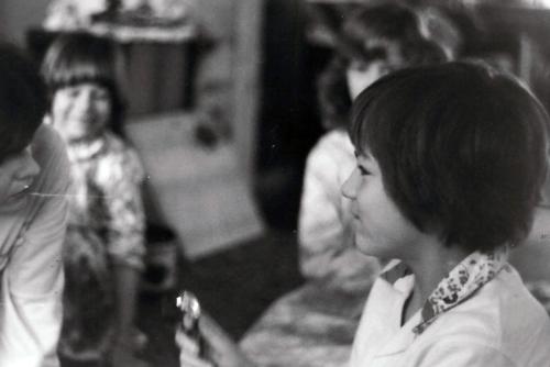 Boys smiles at mother, who is crouched down on the ground to talk to him. Young girls sit in the background. Black-and-white photo.
