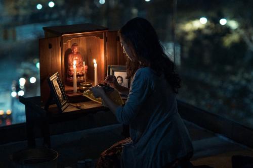 Film still of a woman, kneeling on a rooftop at night, lighting candles in a homemade altar.