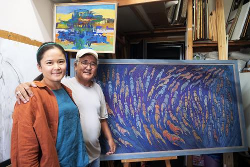 A young woman and older man stand beside a rectangular piece of embroidery on an easel: a herd of orange and blue horses running together, from a high angle, on a blue background.
