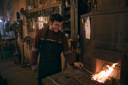 A man holds a metal rod to a red-hot fire in a workshop, smiling.