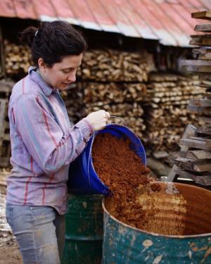 A person pours a bucket full of reddish brown dirt into a large steel drum. In the background are stacks of chopped wood under a metal eave. 