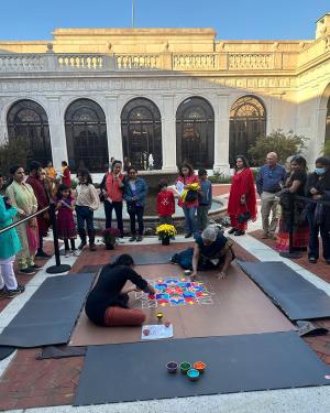 In the brick courtyard of an ornate white stone building, two women sit on the ground, creating a four-sided mandala patterns in lines of white and colored sand. A crowd stands around them.