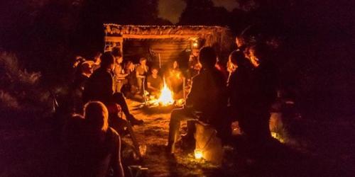 A circle of people are lit in orange by a firepit in the middle.