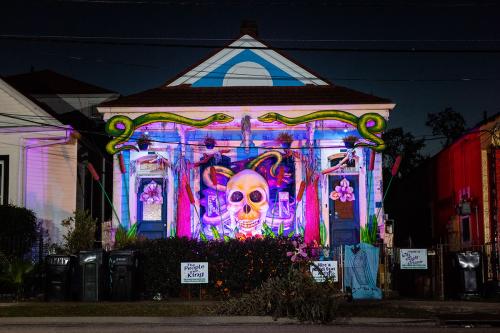 After dark, exterior of a house lit up in purple, with a large human skull sculpture hanging on front. Below the eave of the house are two large painted snakes.