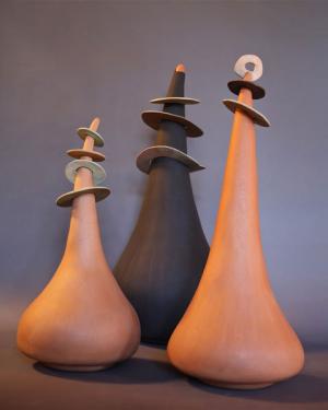 Three teardrop-shaped clay sculptures, with discs around their slender tops, in earthen tones.
