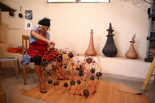 A woman in a red apron sits in her studio working on an abstract wire sculpture. Clay pot-like sculptures are lined up along the wall.