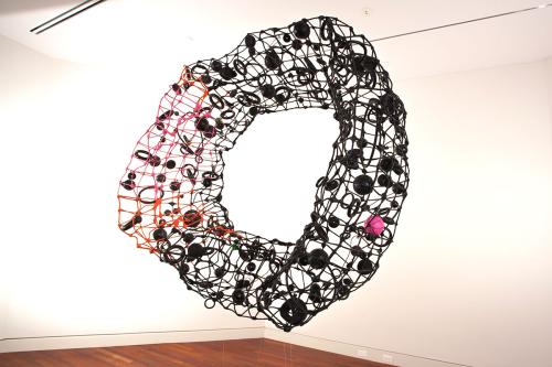 Large sculpture hanging against stark white walls in a gallery. In a ring shape, it resembles a black car tire, made up of pieces of scrap metal and junk.