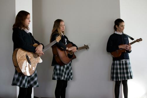 Three young men stand blankly in a hallway, looking like they are waiting for direction. Two hols acoustic guitars and the third a ukulele. They all wear matching school uniforms: navy sweaters, white collared shirts, plaid skirts, and black stockings.