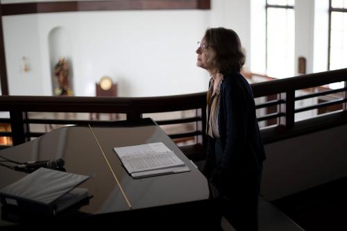 Dramatic shot of an older woman sitting at a piano in the shadows, perhaps in the upper level of a chapel, with sunlight from tall windows brightening the large room behind her.