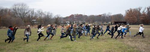Panoramic photo of all the girls of the Oakcrest School Chorus running across an open field, arms splayed out, long hair flying. They are dressed in warm overcoats, scarves and beanies; the bare trees behind them confirm a cold, wintry day.