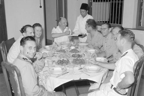A group of white men in light-colored uniforms sit around a dining table set with many dishes. An Indonesian man serves them.