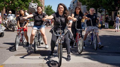 Five young people standing astride lowrider bikes on a town street.