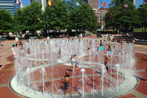 Children play in an outdoor fountain in the shape, from a bird's-eye view, of the Olympic rings. Flags of multiple countries fly above.