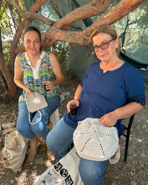 Two women working on crocket projects among low tree branches.