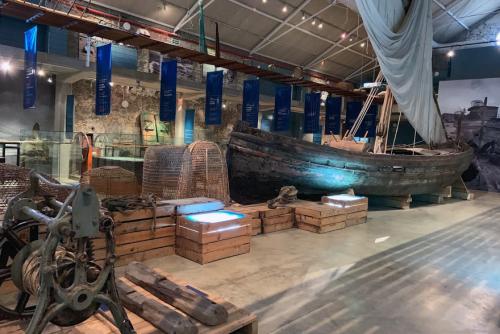 A large, dark room in a museum shows all kinds of fishing gear, woven fish nets, and a wooden boat with canvas mast. Informational screens set in wooden pallet cases are illuminated in blue.
