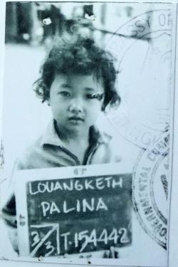 A young girl with short, dark curly hair holds up a handwritten sign with her name: Louangketh Palina, and a series of numbers. On the black-and-white photo is an embassy stamp.