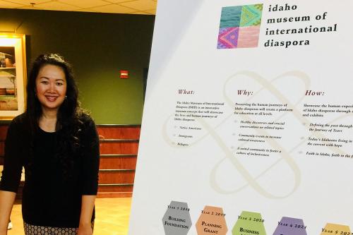 A woman smiles, posing next to a poster introducing the Idaho Museum of International Diaspora.