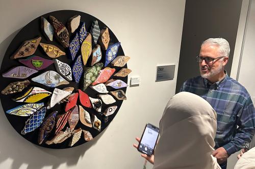 Two people look at a piece of artwork on an office wall, made up of several eye-shaped pieces of palm wood painted and decorated and arranged on a black circular background.
