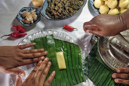 Overhead shot of two pairs of hands making pastels: one person is folding a green banana leaf to create the rectangular pastry. Another person operates a manual press to flatten the balls of dough sitting in a bowl. Other ingredients surround them.