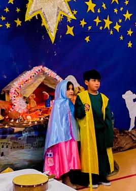 Two young kids dressed in robes stand in front of a nativity scene, with stars hanging above them.