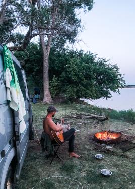 A man strums an acoustic guitar, sitting between a large van and a burning fire pit at the side of a lake.