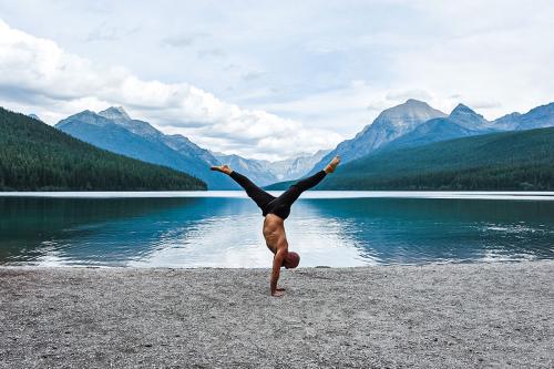 A man, shirtless, does a handstand yoga pose at the edge of a lake, perfectly centered with the mountains in the background.
