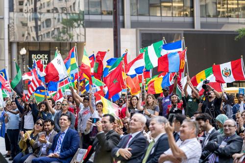A crowd outdoors, half sitting and half standing holding high a variety of national flags.