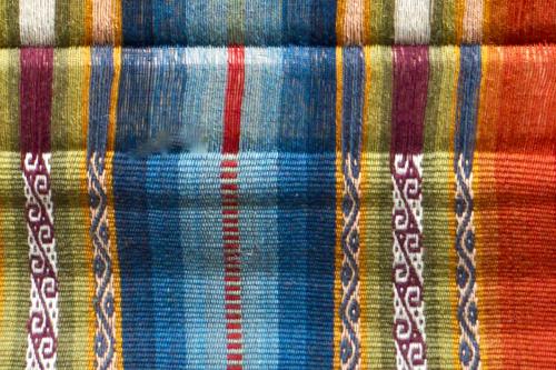 Close-up of an unfinished woven textile in blue, green, orange, and white stripes.