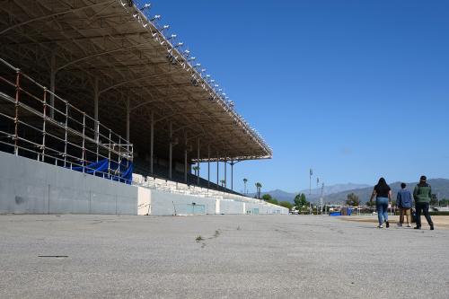 Old stadium seating along a cement track. Three people walk down the track, away from the camera and toward mountains in the distance.