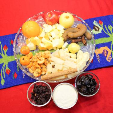From above, bowls of dried hibiscus flowers, white sugar, prunes, and a plate with whole and chopped apples, orange, tamarind, guava, hawthorns, sugarcane, and cinnamon stick.