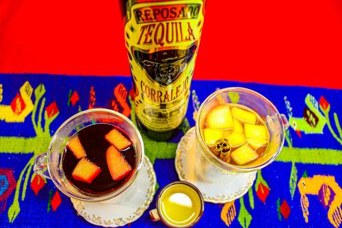 Two glasses of punch with fruit floating in them, plus a cinnamon stick in one, arranged on a tablecloth next to a bottle and shot of tequila.