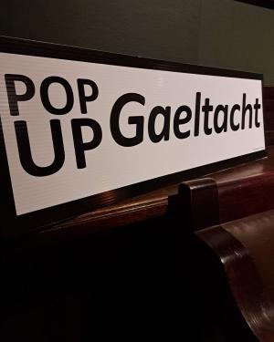 Sign propped up above a hallfway bench that reads Pop Up Gaeltacht.