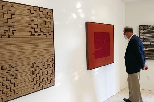 A man examines a red textile art piece in a gallery, with other pieces on either side.