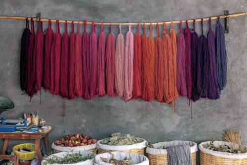 A row of yarn skeins hang from a single wooden pole, arranged in a gradient from deep red to light pink to yellow to purple. Below them, on the ground, are baskets full of dried plant material and other dye sources.