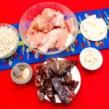 From above, bowls of raw meat, white onion, garlic cloves, dried red chilis, ground spices, and hominy.