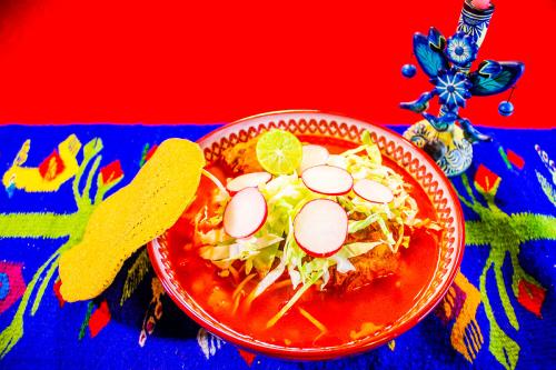In a red bowl, red soup topped with shreed cabbage, lime, and radish slices. Leaning against the bowl is a yellow tostada. Arranged on an embroidered red and blue tablecloth next to a decorative blue candleholder.