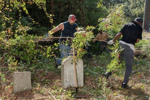 Two men remove fallen branches among gray headstones in a cemetery.