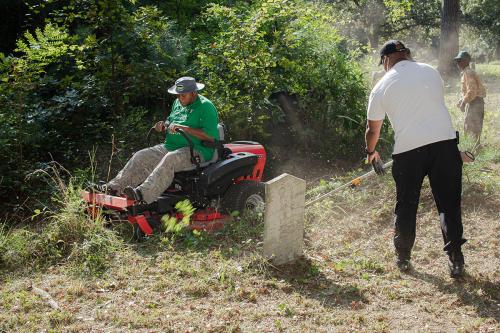 One person uses an edger tool around a gray headstone while another rides a lawn mower through overgrown brush in a cemetery. 