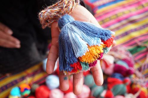 An outstretched hand holds up a tassel made of blue, orange, red, and green yarn. Color balls of yarn and a multicolored striped textile lie below. 