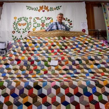 A man unfurls a large quilt with multicolored geometric design.