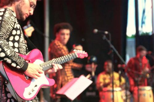 Four people performing on stage. In the foreground, a person on red electric guitar. In the background, blurred, people on various percussion instruments.