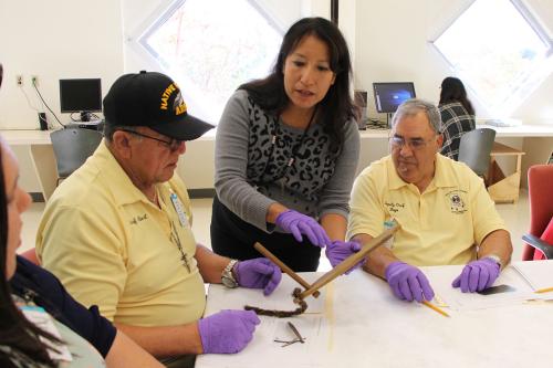 Two men in yellow polo shirts sit at a table with paper and pencils. They direct their attention towards a woman wearing purple nitrile gloves who is holding and gesturing around a wooden tool.