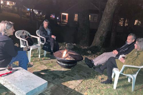 Four people sit, socially distanced, around a metal fire pit at night.
