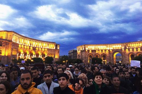 A crowd of people, most with stern expressions, facing forward under a darkening sky. The buildings behind them are lit up.