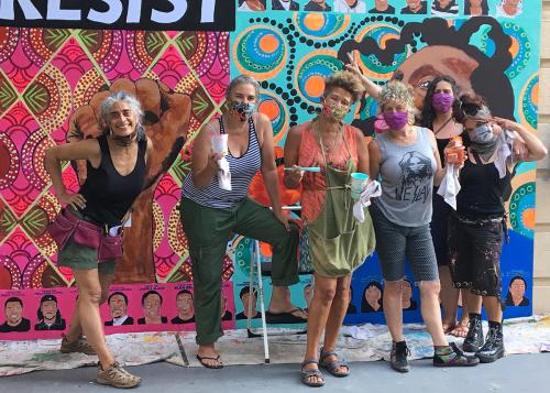 Six people pose in front of a mural with bright patterns in pink, blue, and orange, and the corner of the word RESIST. Some hold paint brushes and cups of paint.