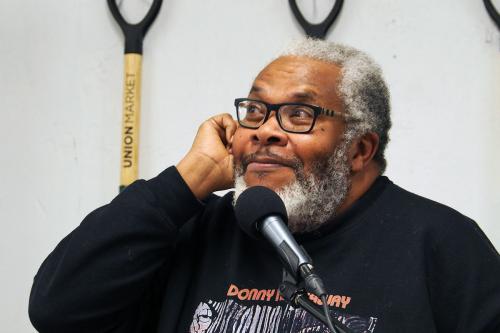 A Black man with graying hair and beard smirks, sitting at a microphone.