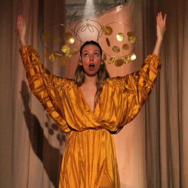 A woman wearing a flowing, shimmery gold dress and a golden mobile-like crown on stage with both arms raised, mouth open. 